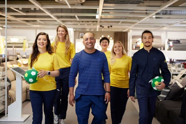 Eine Gruppe von IKEA Mitarbeiter:innen mit gelben und blauen Uniformen lächelt in die Kamera.