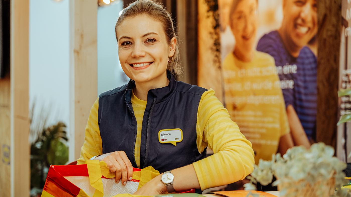 Eine IKEA Mitarbeitender in gelber IKEA Uniform unterhält sich lachend mit IKEA Kund:innen mit Einkaufswagen.