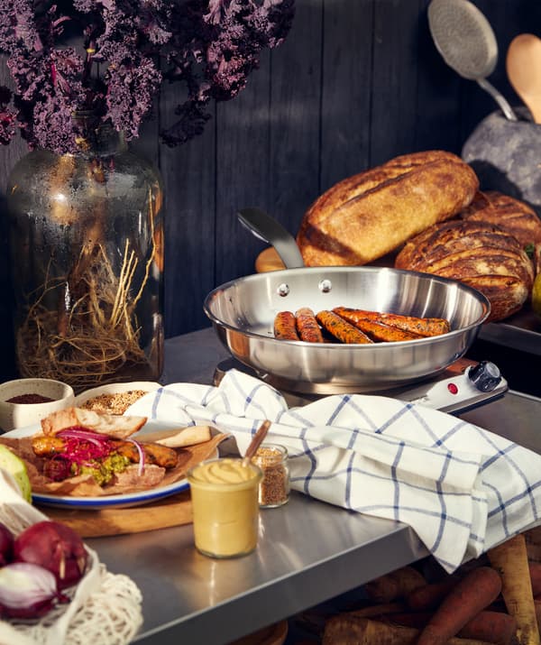 Eine Metallarbeitsfläche, u. a. mit Würstchen in einer SENSUELL Bratpfanne aus Edelstahl, einem Teller mit Essen und mehreren Laiben Brot.
