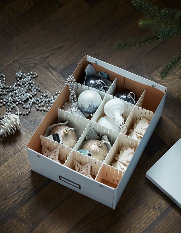 Eine ordentlich aufgeräumte Aufbewahrungsbox auf einem Holzboden enthält verschiedene Weihnachtsschmuckstücke, darunter silberne und goldene Kugeln, die für eine festliche und aufgeräumte Atmosphäre sorgen.