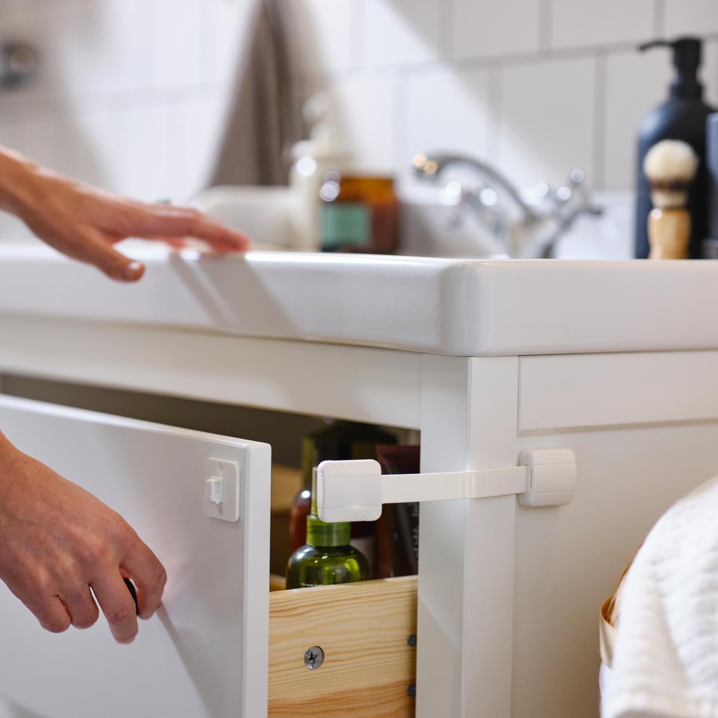 Eine Person hält mit der Hand eine Schublade eines weißen HEMNES Waschbeckenschranks offen, an der eine weiße UNDVIKA Multisperre angebracht ist.