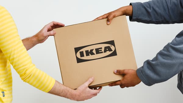 Eine Person in einem grauen Pullover reicht einen Karton mit schwarzem IKEA Logo einer Person in einem gelben IKEA Mitarbeiteroberteil.