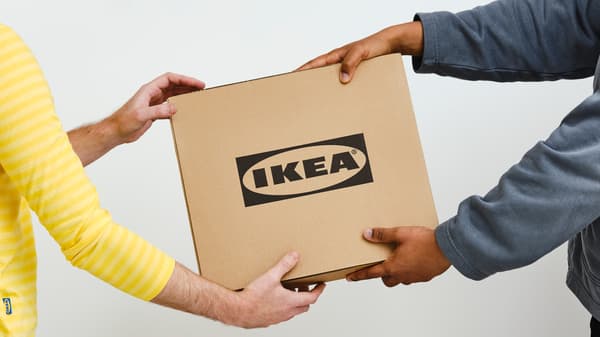 Eine Person in einem grauen Pullover reicht einen Karton mit schwarzem IKEA Logo einer Person in einem gelben IKEA Mitarbeiteroberteil.