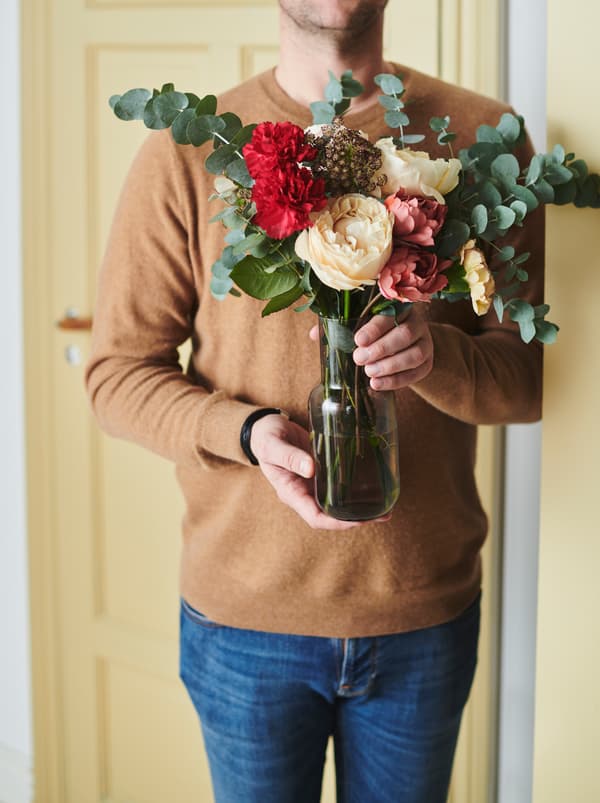 Eine Person in Jeans und einem Pullover, die eine FÖRENLIG Vase mit SMYCKA Kunstblumen in den Händen hält.