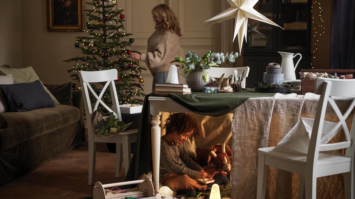 Eine Person schmückt den Weihnachtsbaum und unter dem Tisch im Vordergrund spielt ein Kind halbversteckt mit Spielzeug.