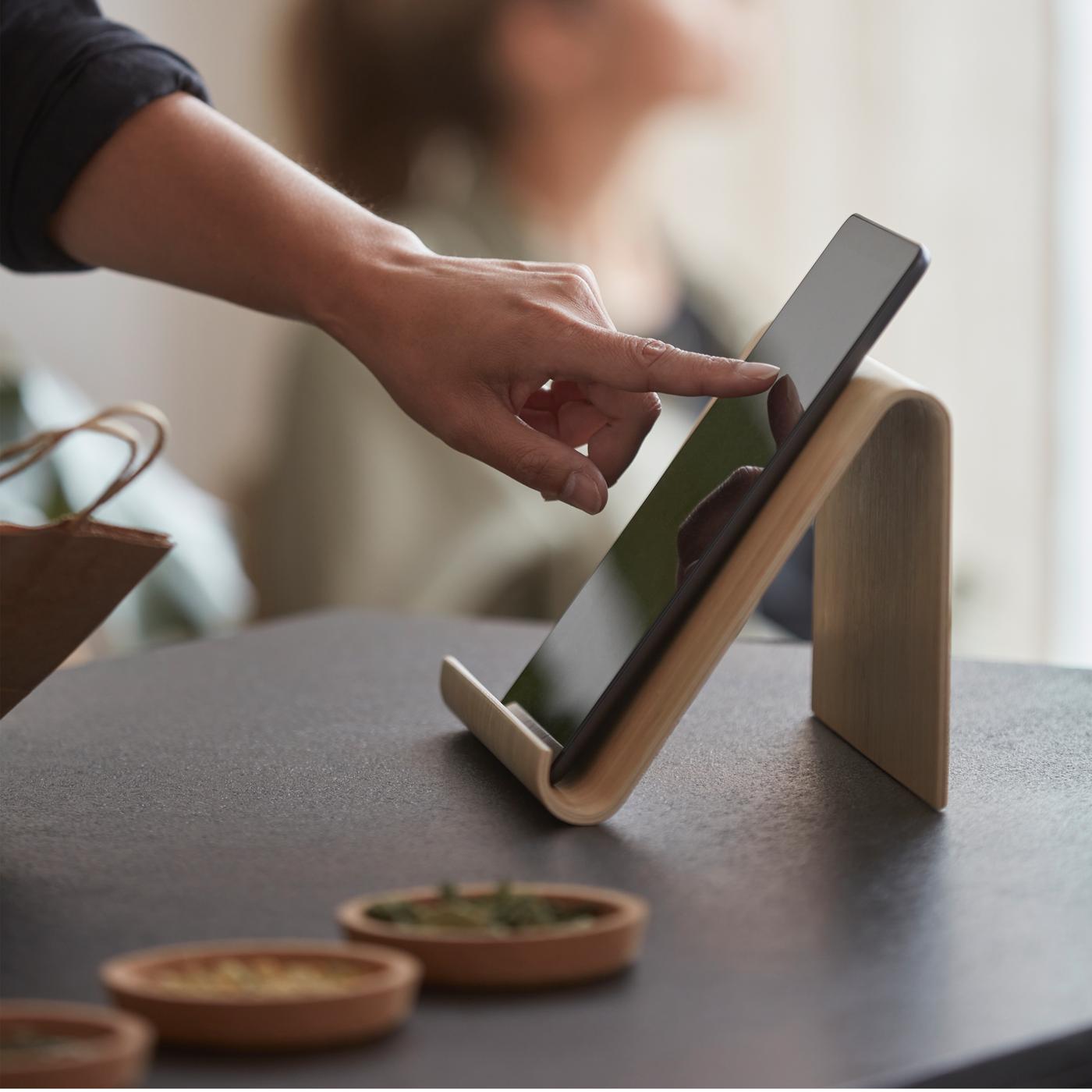 Eine Person scrollt auf einem Tablet, das auf einem Tablet-Halter aus Bambus auf einer schwarzen Arbeitsplatte steht. Eine weitere Person sitzt im Hintergrund am Fenster.