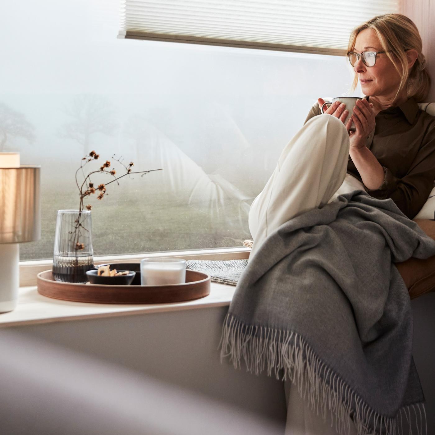 Eine Person sitzt am Fenster und geniesst eine Pause bei einer Tasse Tee. Auf der Fensterbank steht eine SYMFONISK Tischleuchte mit WiFi-Speaker.