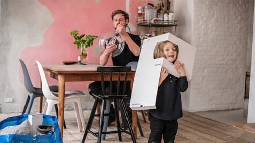 Eine Person sitzt an einem Esstisch und montiert eine Leuchte, während ein Kind mit der Pappverpackung spielt.