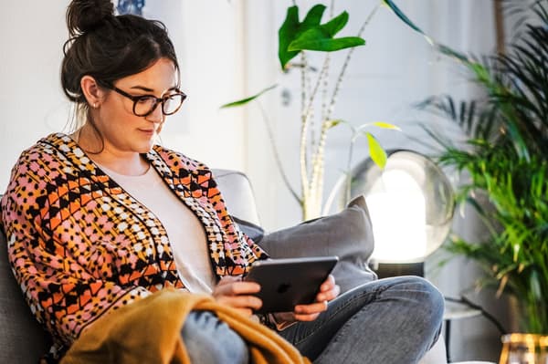 Eine Person sitzt auf dem Sofa und hält ein Tablet in der Hand.