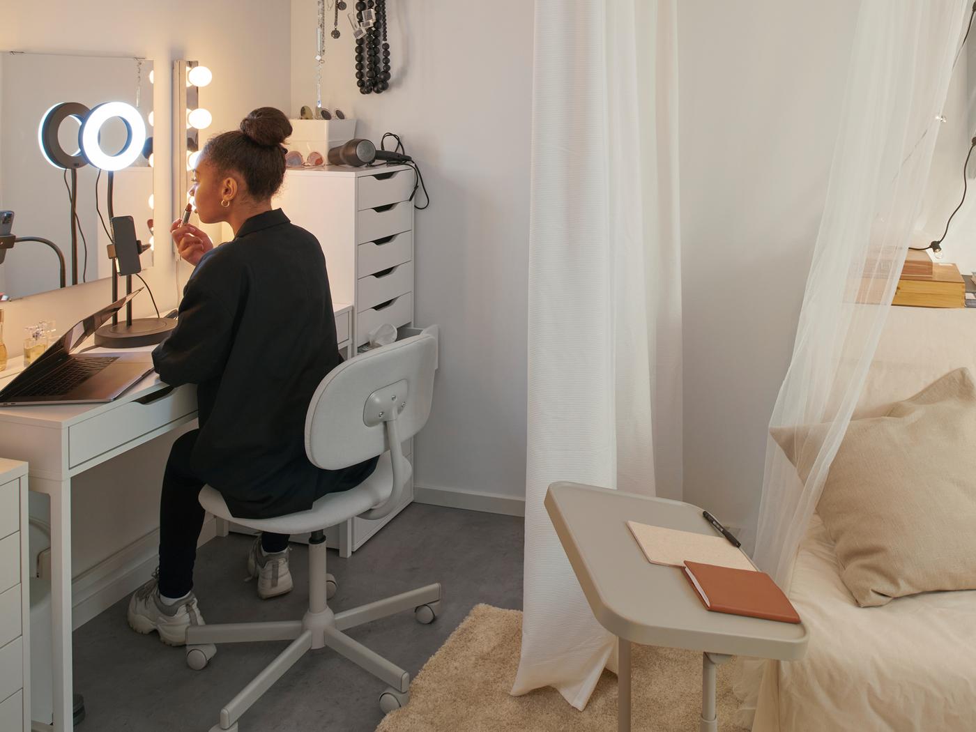 Eine Studentin mit langen Haaren sitzt in einem Wohnheimzimmer auf einem BLECKBERGET Drehstuhl an einem MICKE Schreibtisch. Im Licht einer LÅNESPELARE Ringleuchte mit Mobiltelefonhalter trägt sie Lippenstift auf.