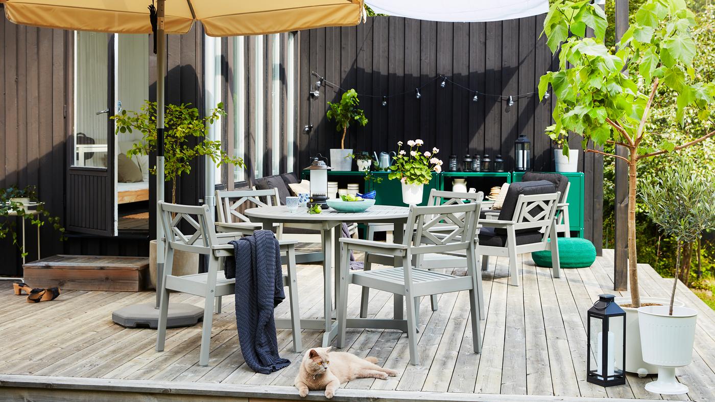 Eine Terrasse mit Holzboden, einem beigen Sonnenschirm und grauen Möbeln für aussen. Vor der Sitzgruppe liegt eine rote Katze.