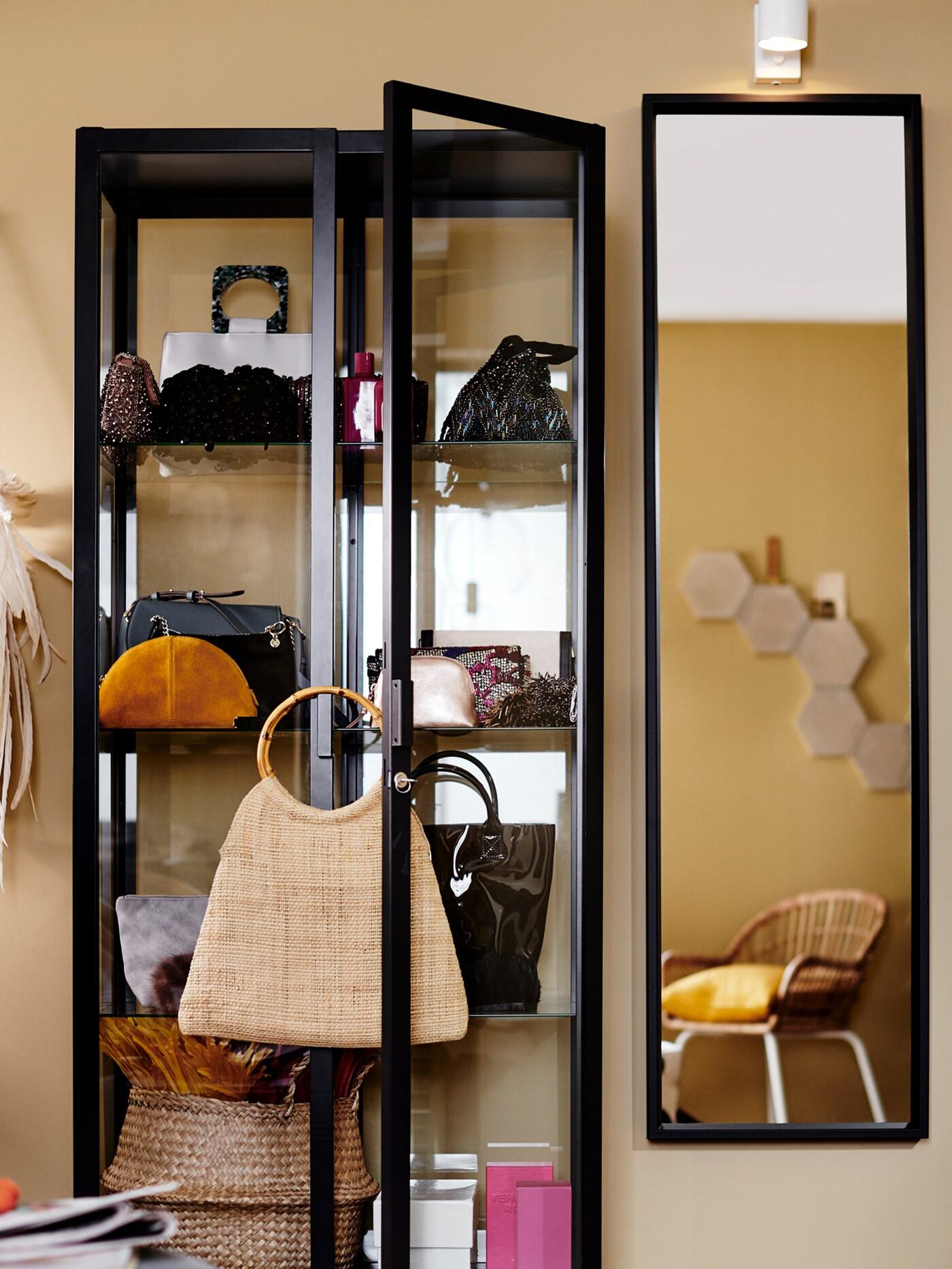 Eine Vitrine mit einer Vielzahl verschiedener Gegenstände, u. a. Handtaschen, steht neben einem Spiegel an einer Wand.