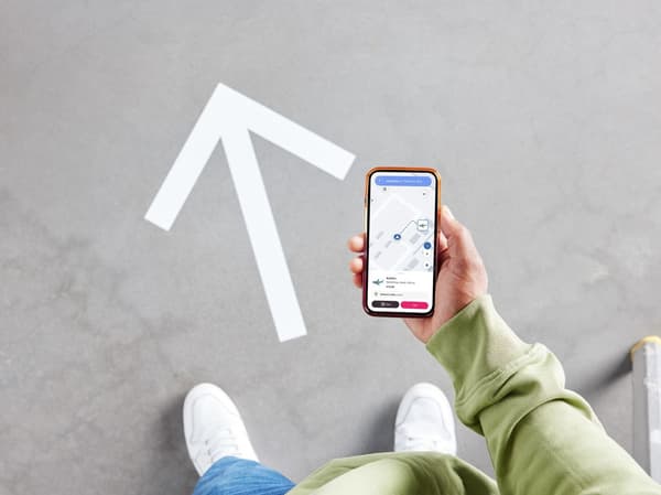 Eine von oben gesehene Person steht in einem Einrichtungshaus neben einem grossen, auf dem Boden gedruckten Pfeil. Die Person hält ein Smartphone, auf dem ein Plan der Abteilung angezeigt wird.