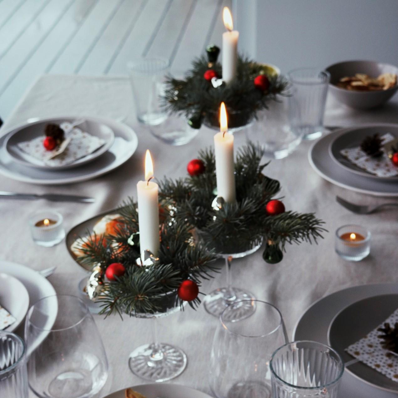 Elegant holiday table setting with lit candles on pine wreaths, white plates, and clear glasses. Festive, warm ambiance with red and green decor.