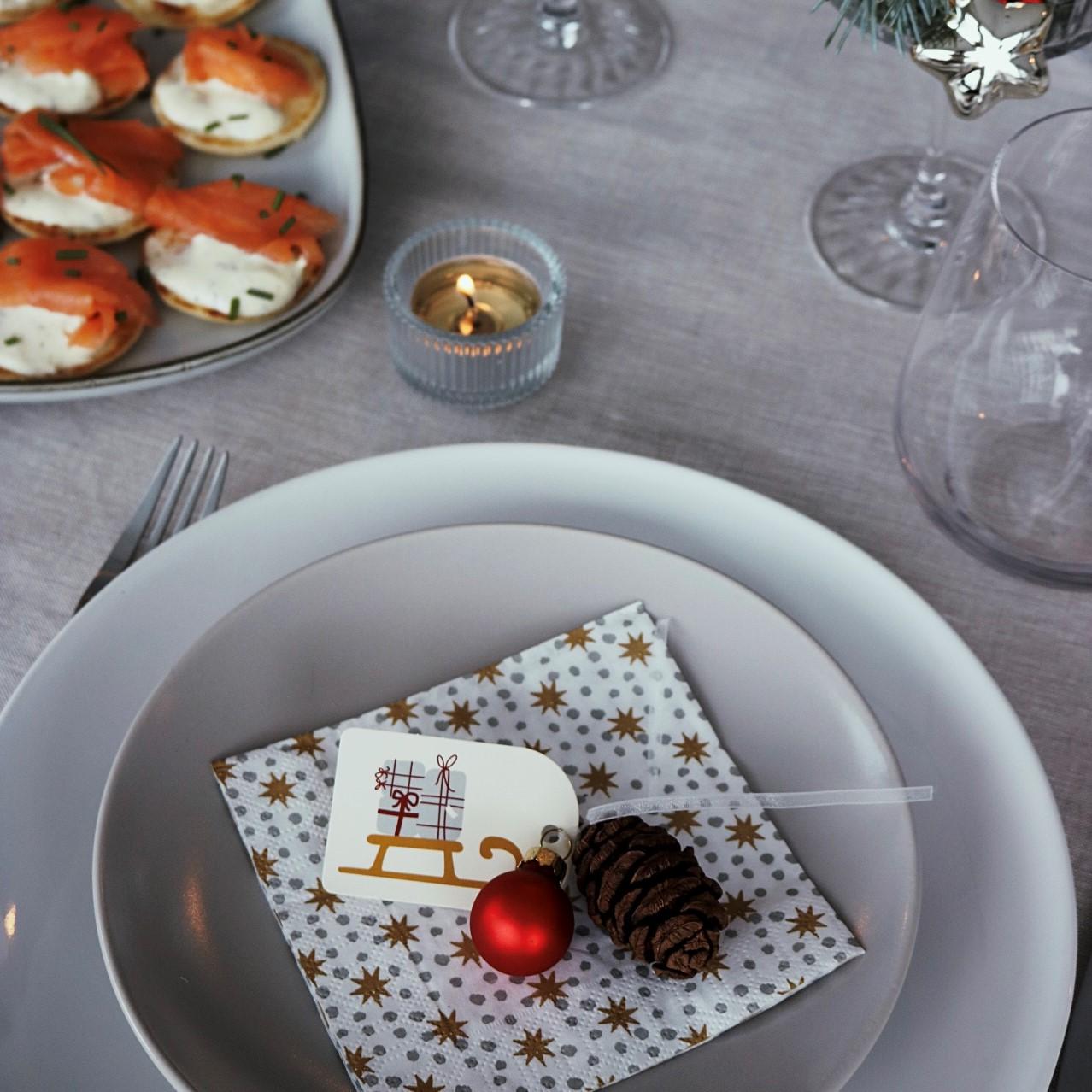 Elegante tavola natalizia con piatto bianco, tovagliolo festivo, ornamento rosso, pigna e biglietto decorativo. Accanto ci sono antipasti di salmone affumicato e una candela accesa.