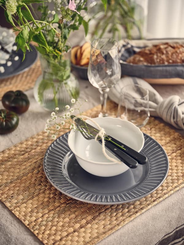 Elegante Tischdekoration mit einer weissen Schale auf einem grauen Teller, gebundenen Besteckteilen und Blumen, auf einer strukturierten Tischmatte. Brot, Gläser und Blumen schaffen eine gemütliche Atmosphäre.