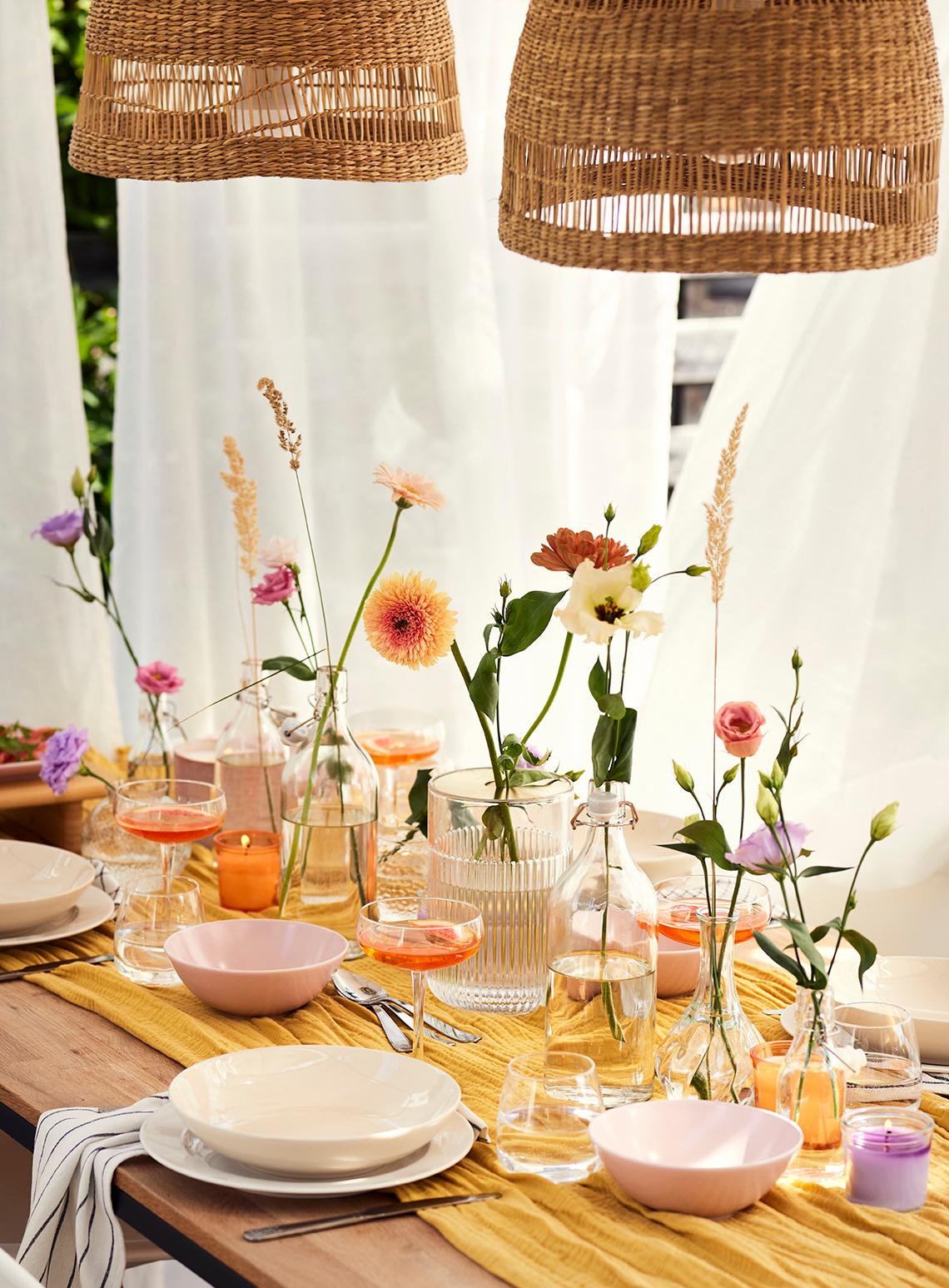 Eleganter Esstisch im Freien mit geflochtenen Lampenschirmen, pastellfarbenem Geschirr und frischen Blumen in einer Gartenlandschaft.