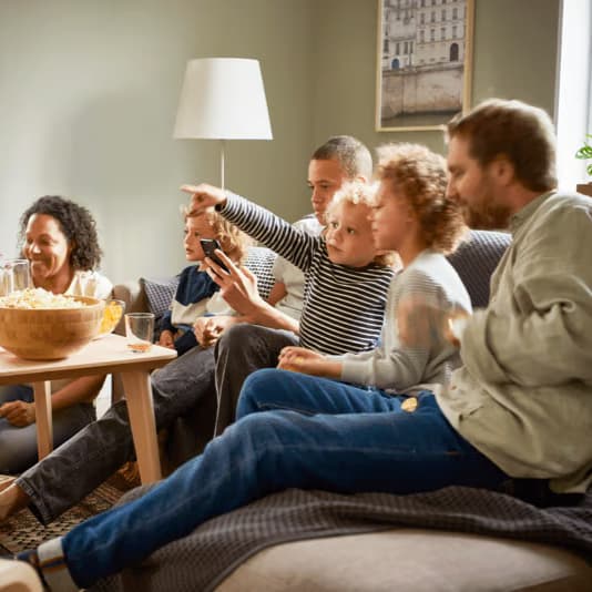 En grupp personer sitter tillsammans i ett vardagsrum, avslappnat samlade i soffor och fåtöljer. De pratar och gestikulerar medan de delar snacks, bland annat en stor skål popcorn på ett träbord framför dem. Rummet är hemtrevligt inrett med en golvlampa och en tavla på väggen.