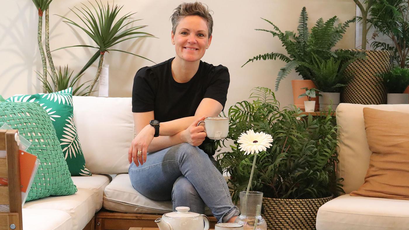 En kvinde sidder i en NÄMMARÖ modulopbygget havesofa med hvide hynder og grønne puder omgivet af grønne planter.