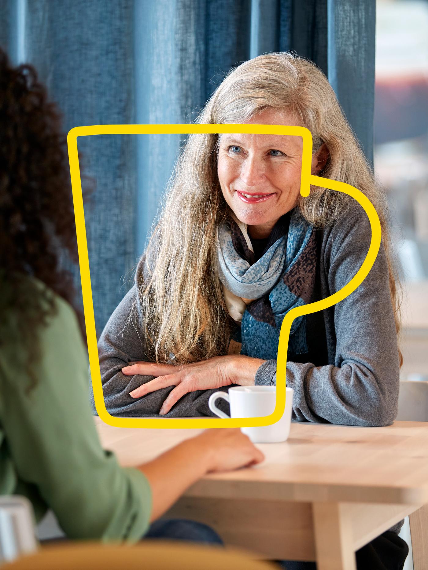 En kvinde sidder og smiler ved et bord med en kop kaffe foran sig