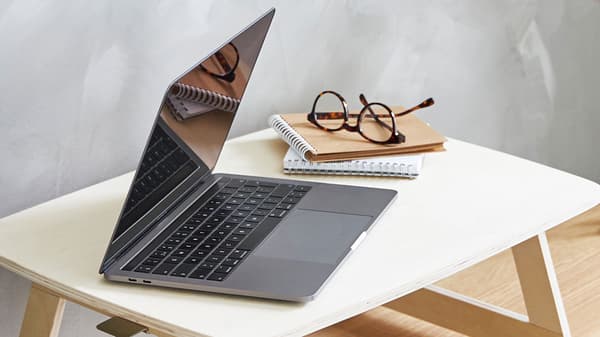 En öppen laptop på ett laptopbord i ljust trä, med ett par glasögon och anteckningsblock bredvid. 