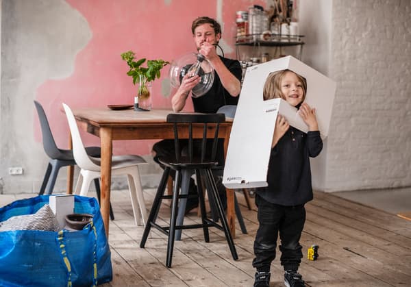 En pappa sitter vid matbordet och monterar en lampa medans hans son leker med kartongen. 