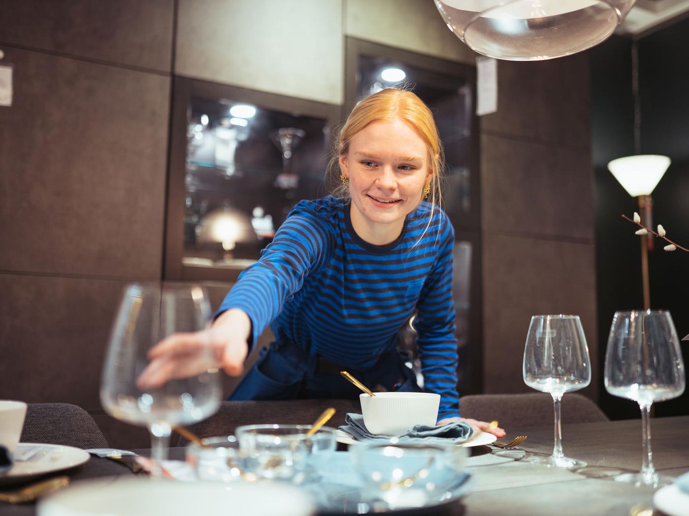 En person med lyst hår iført en blå og sort stribet skjorte ses dække et bord, der inkarnerer værdien af en elev i IKEA. Bordet er dækket med vinglas, skåle, tallerkener og bestik. Baggrunden har et moderne interiør med mørke skabe og en gulvlampe.