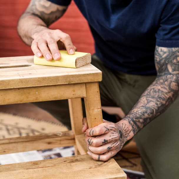 En person med tatueringar på armarna använder sandpapper för att slipa en pall i trä.