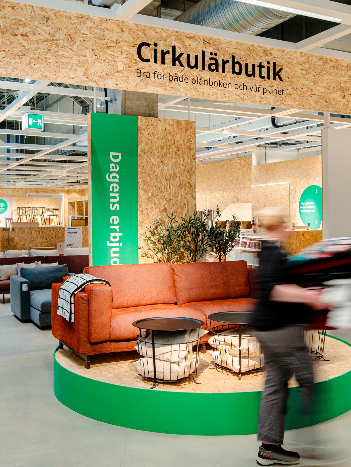 En röd soffa med små svarta bord framför och en person i svart tröja som bär på en möbel går förbi. Ovanför står det Cirkulärbutik på en skylt. 