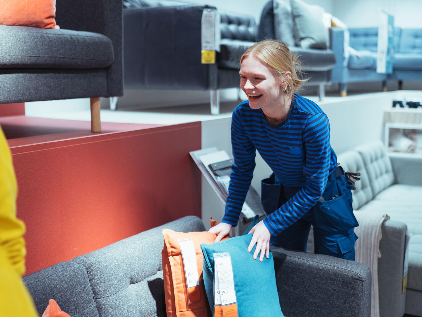 En smilende person i en blå- og sortstribet skjorte arrangerer blå og orange puder på en grå sofa i en møbelbutik, der inkarnerer værdien af kundepleje. Forskellige sofaer og møbler er udstillet rundt om i butikken, der minder om en elev i IKEA, der finpudser sine færdigheder.