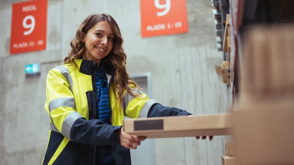 En smilende person med langt hår i en synlig jakke er ved at organisere kasser på en hylde i et lager. To store røde skilte med tallet 9 er synlige i baggrunden, der viser muligheder for en elev i IKEA.