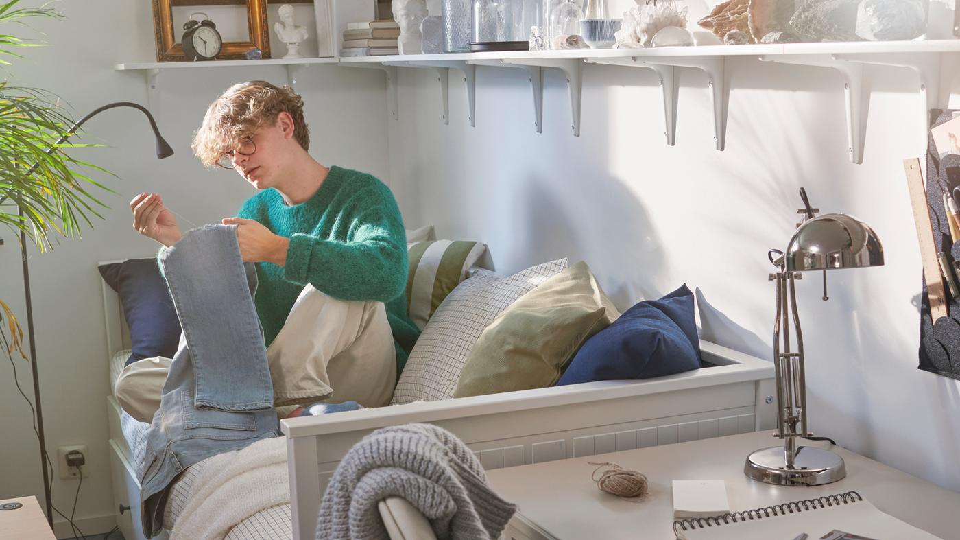 En studerende sidder på en HEMNES sovesofa og syr et par jeans. På hylderne på væggen står der et vækkeur, rammer og genstande.