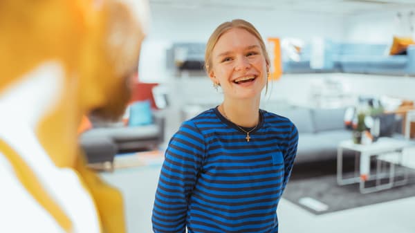 En ung person med lyst hår smiler bredt, mens hun står indendørs. Hun er iført en blå og sort stribet skjorte. Baggrunden viser et moderne, stærkt oplyst rum med møbler, og en sløret figur er synlig i forgrunden, der minder om den imødekommende atmosfære på ledertrainee forløbet i IKEA.