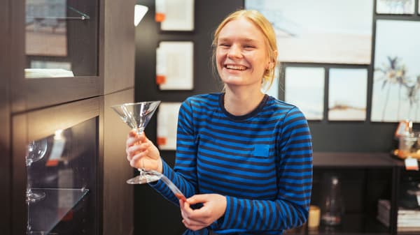En ung person med lyst hår smiler og holder et martini-glas i en moderne, veloplyst butik. De er iført en blåstribet skjorte og står ved siden af glashylder med indrammede billeder synlige på væggen bag dem, der inkarnerer værdierne af en elev i IKEA.
