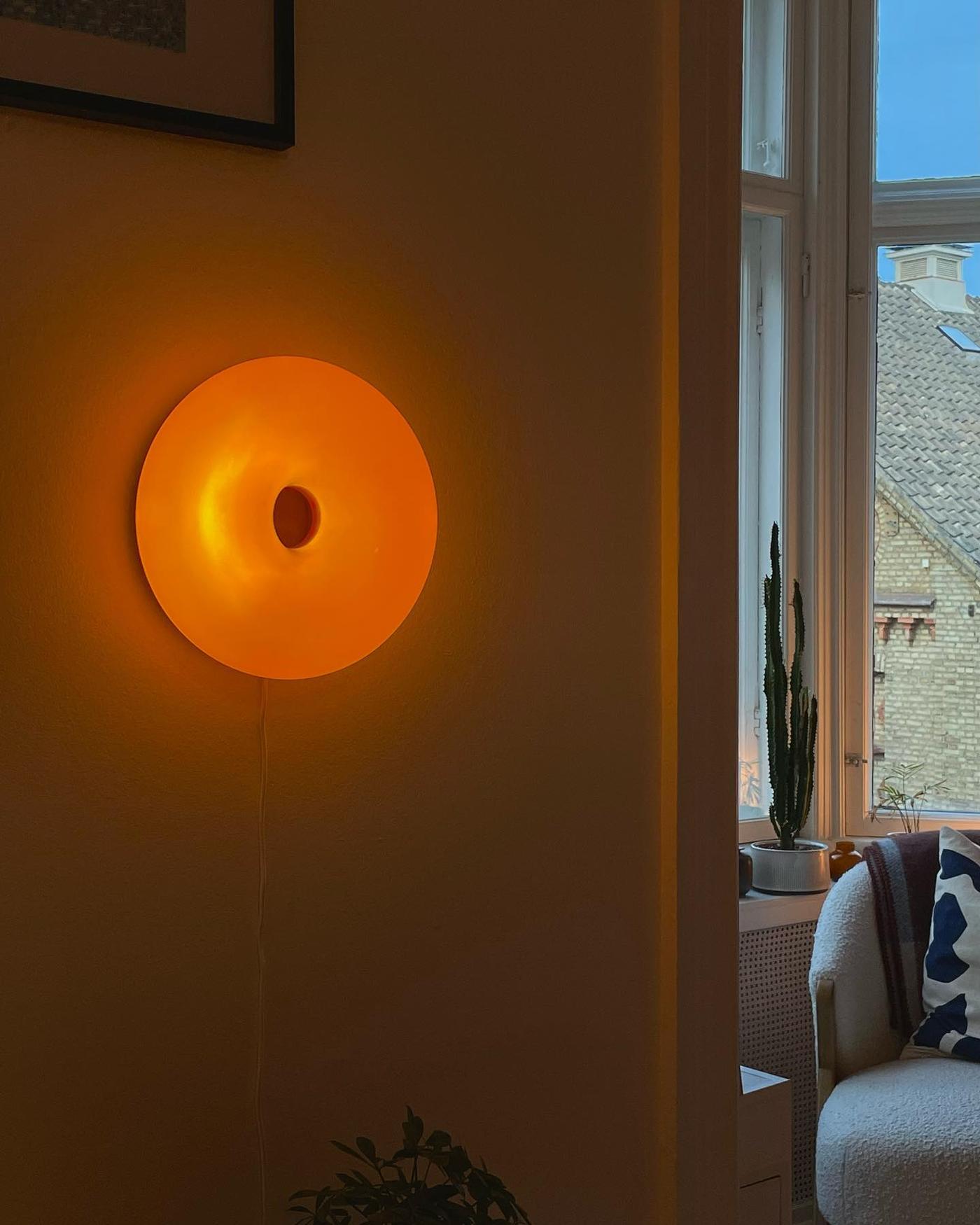 En VARMBLIXT vägglampa lyser varmt orange på en vägg i ett rum där ett fönster och en fåtölj syns i bakgrunden.