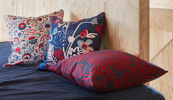 End or side of a made bed by a plywood-panelled wall. Three richly patterned cushions decorate the bed.