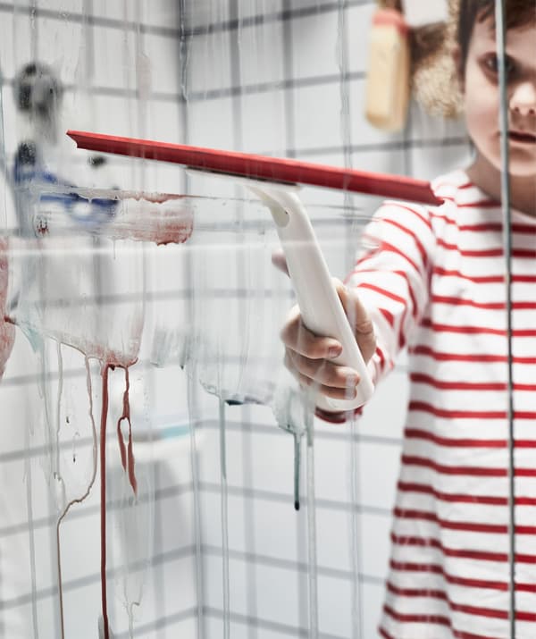 Enfant en chemise rayée debout dans la douche en train de manipuler une raclette pour enlever la peinture à l’aquarelle de la cloison en verre.