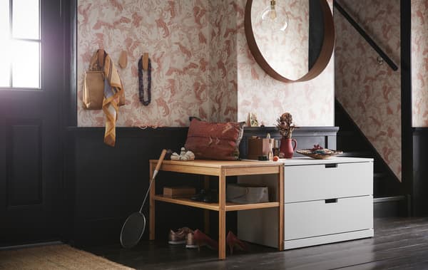 Entre med en lysegrå NORDLI kommode med 2 skuffer under et rundt spejl og ved siden af en bænk af træ med åbne hylder.