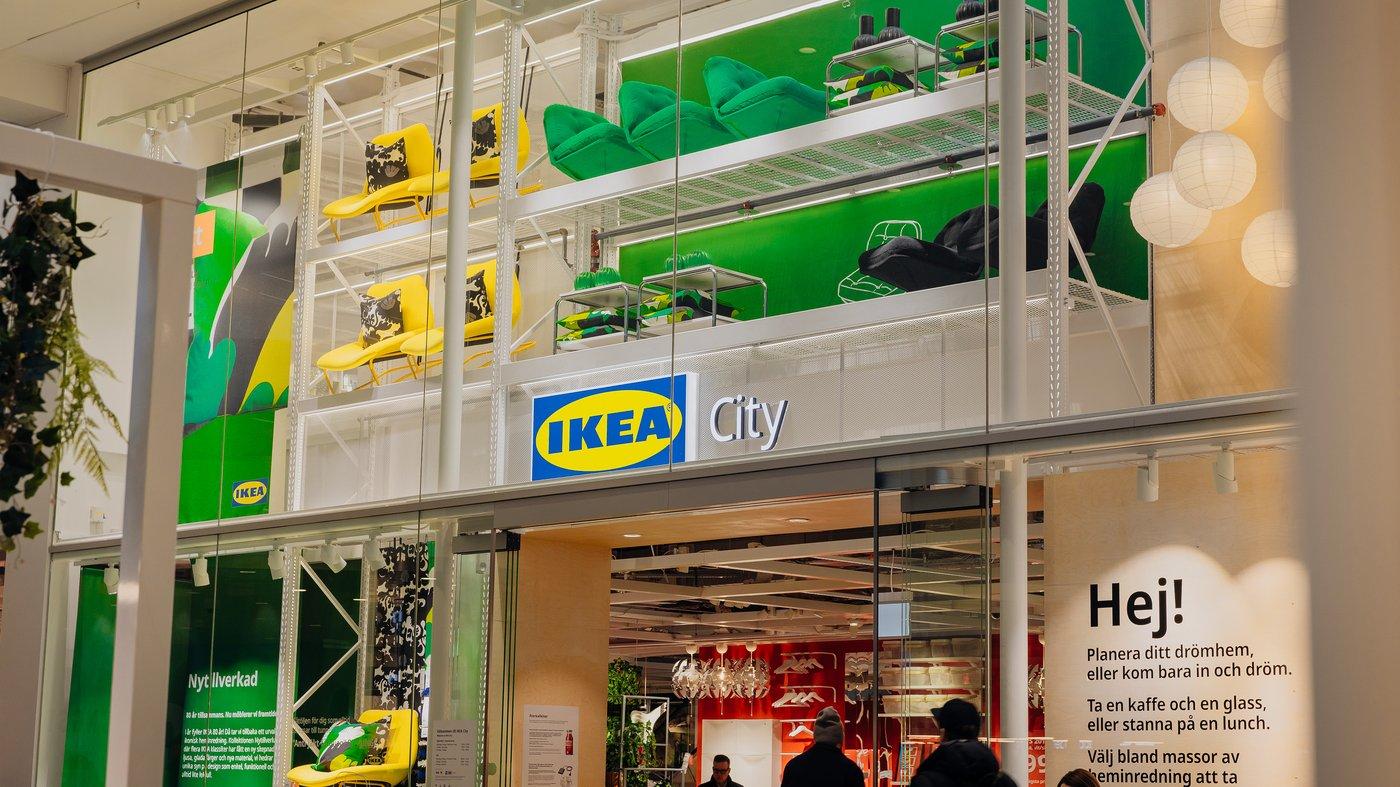 Entrén till en IKEA City Gallerian med stora glasfönster. Ovanför ingången syns hyllor med färgglada fåtöljer och kuddar i grönt, gult och svart. Inne i butiken syns belysning och dekorationer samt några personer som rör sig i lokalen.