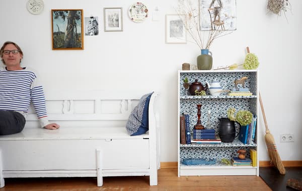 Erik sitting on a white bench against a white wall, next to a floral patterned bookcase filled with books and ornaments.
