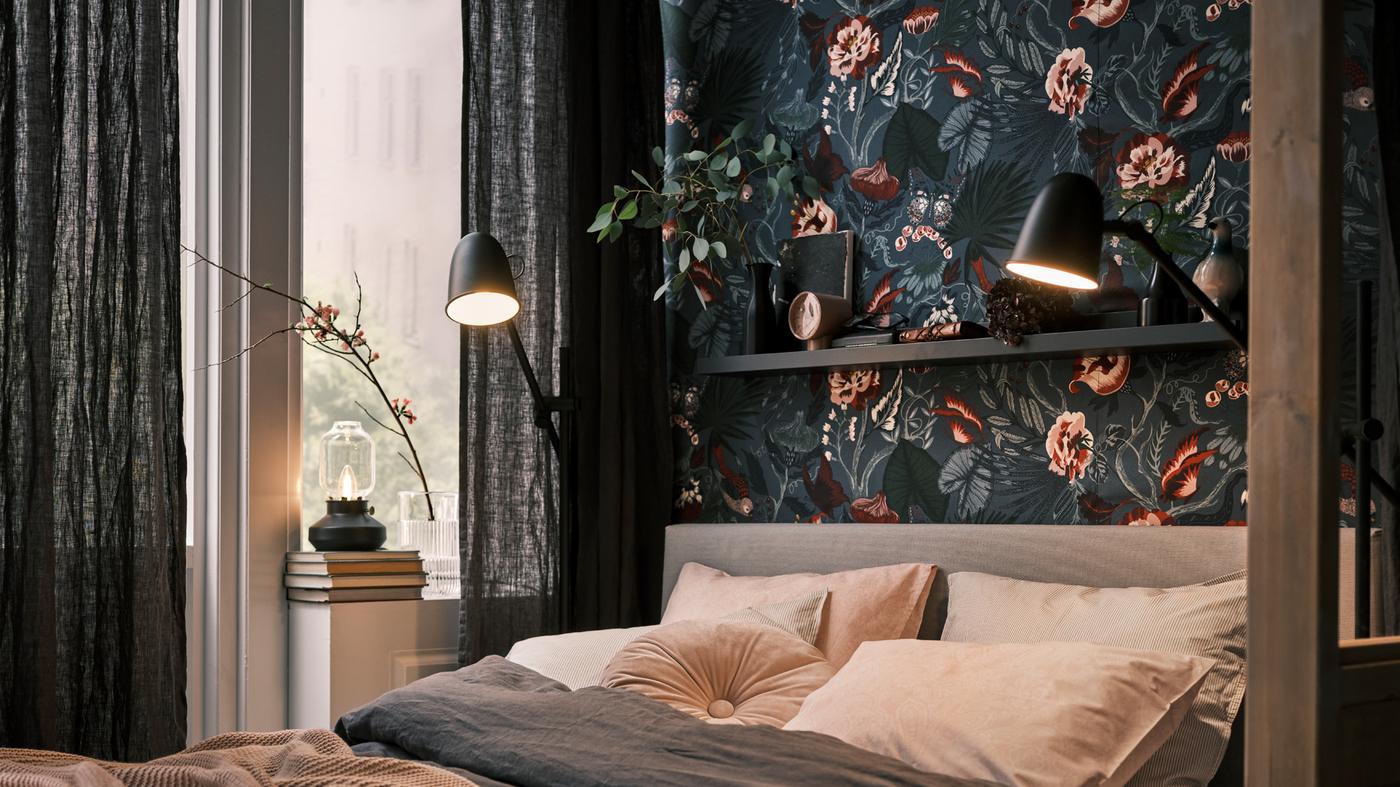 Es ist ein Bett vor einer dunklen Wand mit Blumenmuster zu sehen. Rechts und links davon befinden sich jeweils schwarze SKURUP Stand-/Leseleuchten. Auf der Fensterbank steht noch eine Tischleuchte.