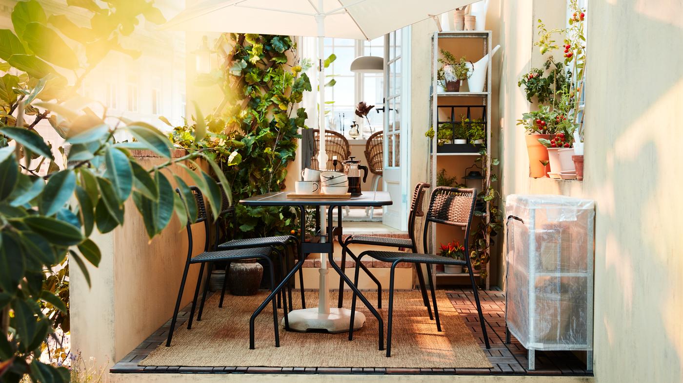 Es ist ein grüner Balkon mit einem schwarzen Essplatz-Set, einem weissen Sonnenschirm, einem gewebten Teppich und vielen Pflanzen zu sehen.