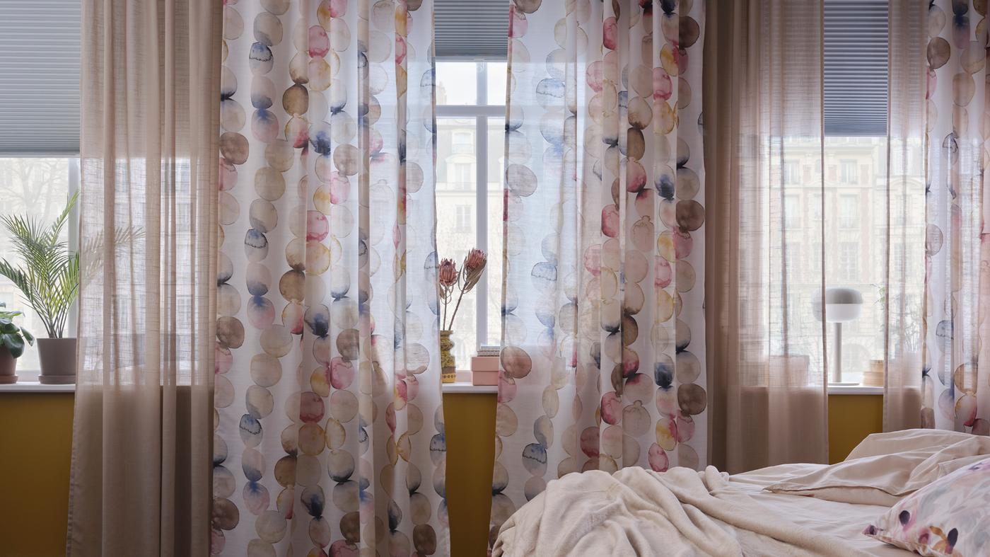 Es ist ein Schlafzimmer mit einer Reihe von Fenstern, bunten SÄCKMAL Gardinen, beigefarbenen Gardinenstores und weißen verdunkelnden Faltjalousie zu sehen.