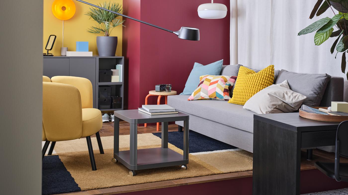 Es ist ein Wohnzimmer im Retrostil mit einem grossen grauen ÄLVDALEN Bettsofa, einem gelben Sessel und einem grauen Couchtisch zu sehen. Die Wände sind in kräftigen Farben gehalten.