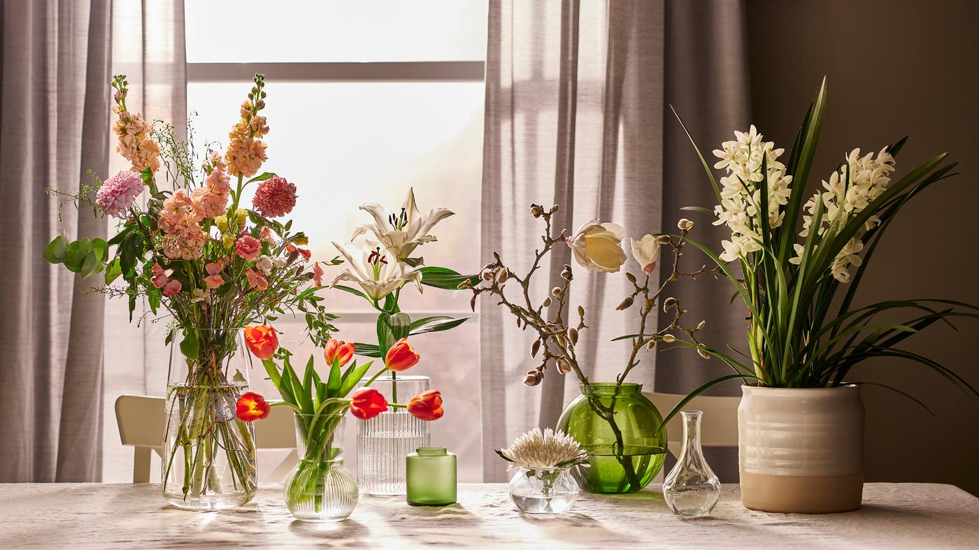 Et bord ved vinduet med forskellige blomsterarrangementer i grønne glasvaser og en råhvid stentøjsvase.