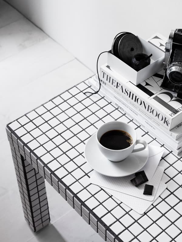 Ett bord med svartvitt rutmönster. På bordet står en vit kopp med svart kaffe på ett vitt fat. Bredvid ligger två små chokladbitar på en vit servett. I bakgrunden till höger syns en stapel med böcker.