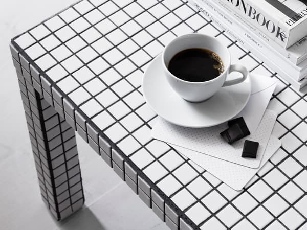 Ett bord med svartvitt rutmönster. På bordet står en vit kopp med svart kaffe på ett vitt fat. Bredvid ligger två små chokladbitar på en vit servett. I bakgrunden till höger syns en stapel med böcker.