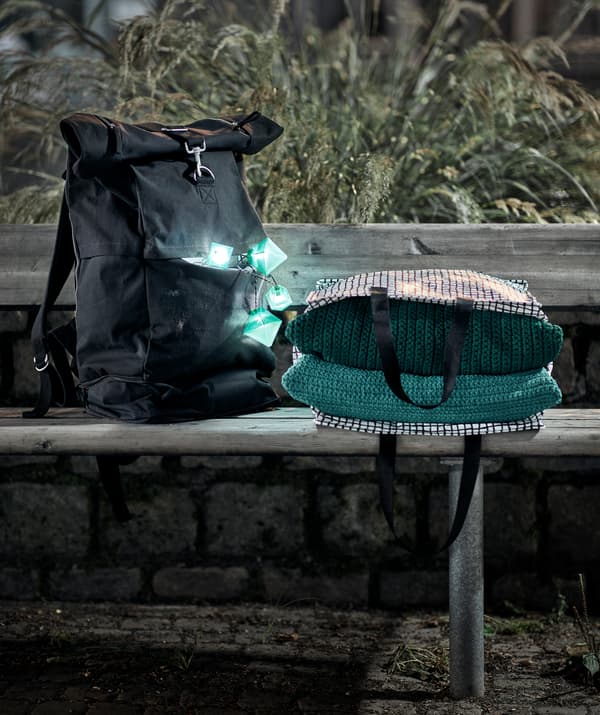 Evening setting with a public bench on which are placed a backpack, lit fairylights in its pocket, and a bag with textiles.