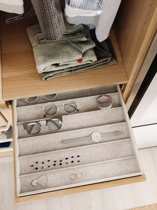 Eyewear, a watch and jewelry in neat rows in a KOMPLEMENT insert within a pulled-out, shallow drawer in an open wardrobe.