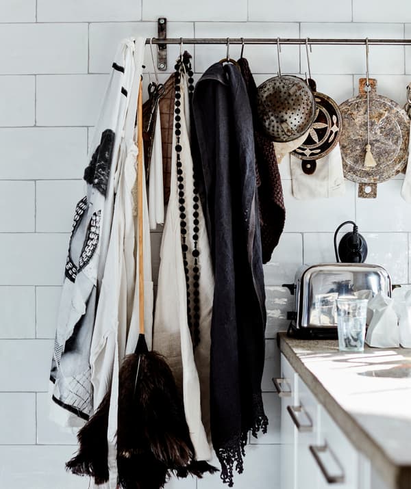 Fabric, a duster and colanders hanging on a metal rail on a white tiled wall.